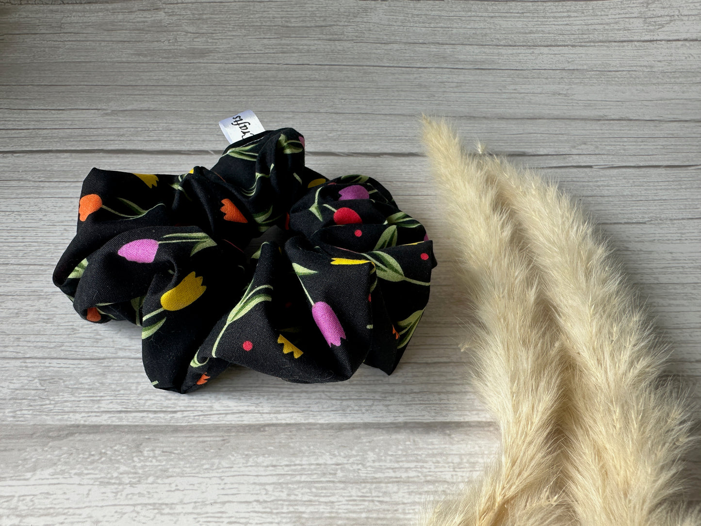 The Cotton Extra Large Scrunchie - Tulips by Crafts by Kate features a luxurious design with colorful floral patterns, lying on a wooden surface next to beige pampas grass.