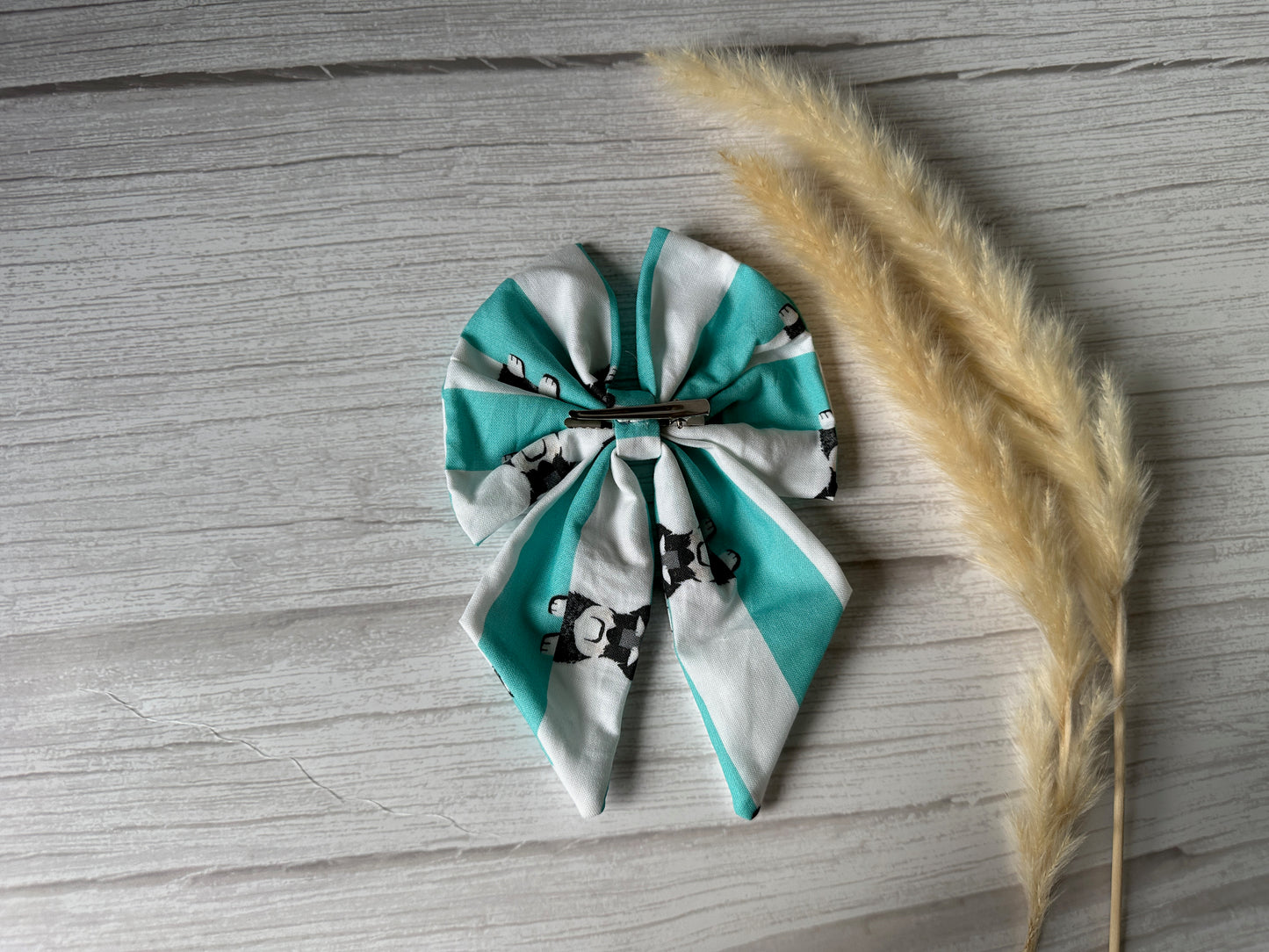 A Cotton Sailor Bow Clip - Striped Schnauzers by Crafts by Kate lies on light wood, exuding handcrafted luxury, with teal and white panda patterns beside dried pampas grass.