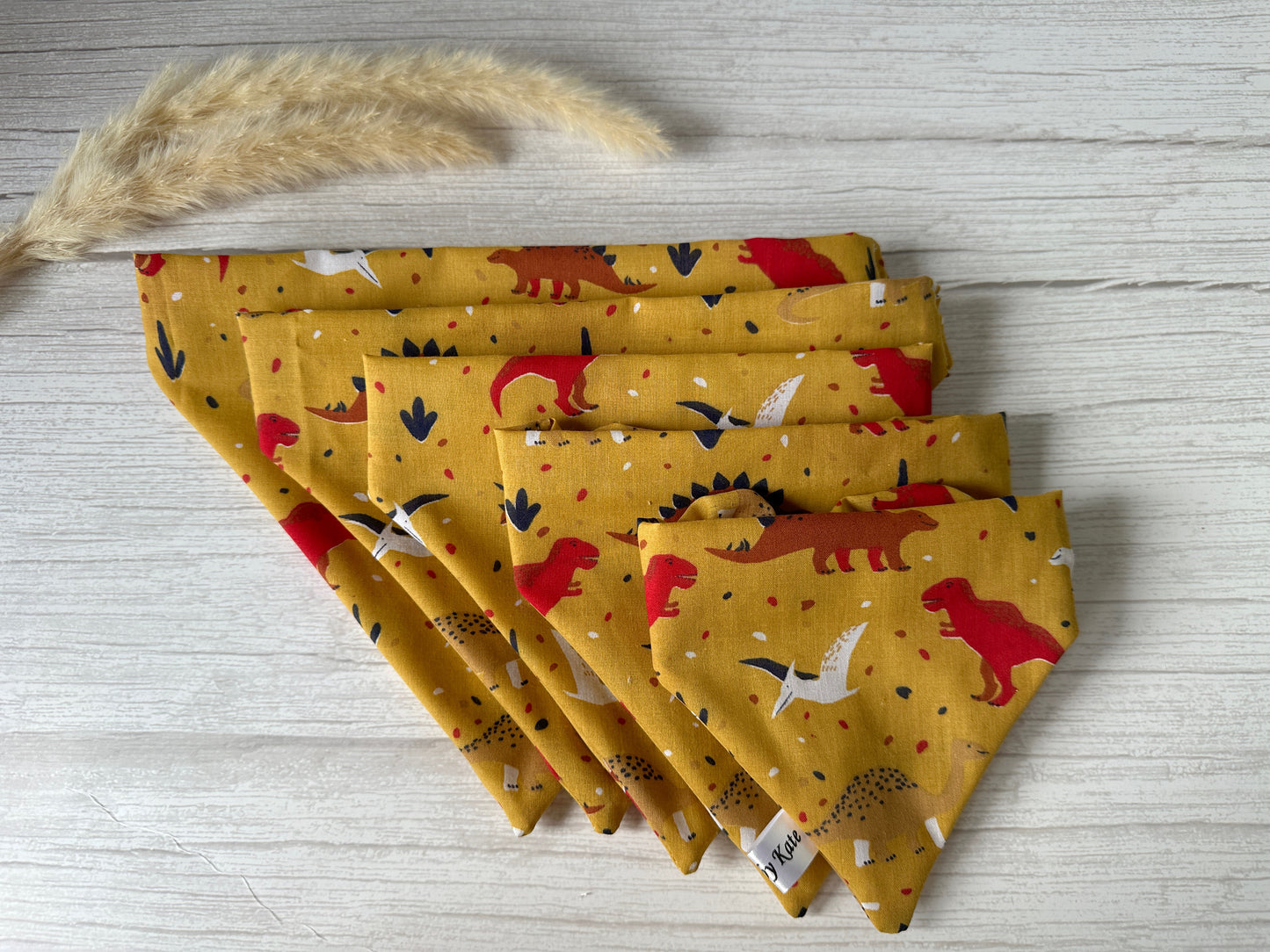 Five yellow napkins featuring the colorful Dinosaur Lovers Cotton Pet Scrunchie Bandana pattern by Crafts by Kate are spread out on a light wooden surface. The napkins are arranged in descending order of size from left to right. A beige, fuzzy plant stem lies next to the smallest napkin, making it an ideal setup for any dinosaur enthusiast or pet bandana project.