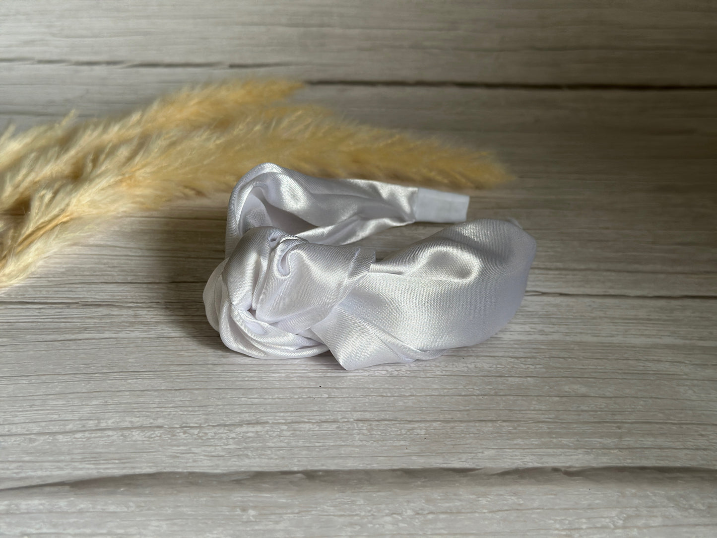 The "Silk Knotted Headband - Ice White" by Crafts by Kate elegantly rests on a textured wooden surface, showcasing its luxurious knot design. In the background, delicate strands of beige pampas grass enhance the scene with an elegant touch.