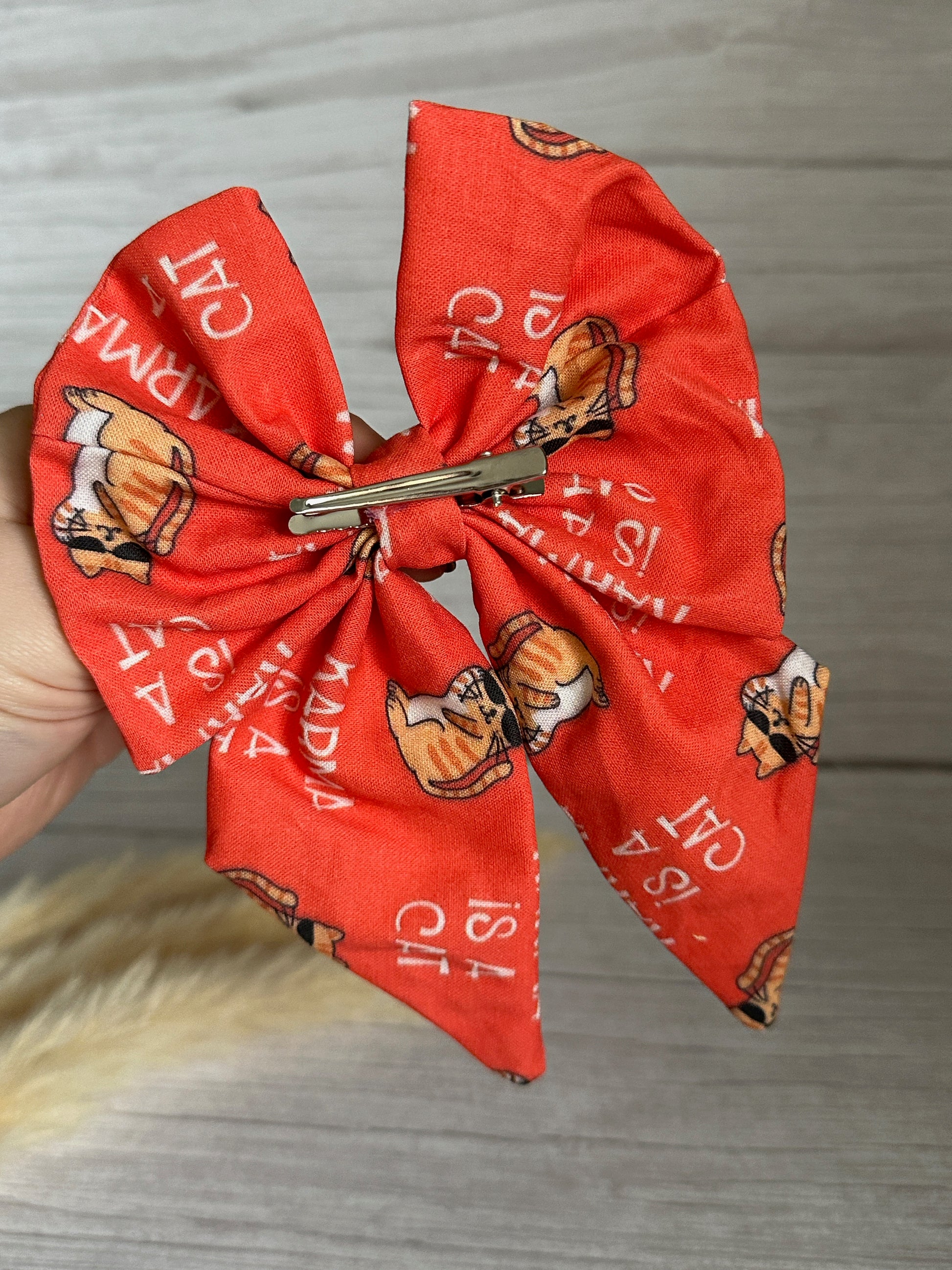 A hand holding the luxurious "Cotton Sailor Bow Clip - Karma is a Cat" by Crafts by Kate. The red cotton bow features a charming pattern of an illustrated dog wearing glasses and a scarf, along with white text in Italian. A silver clip is attached to the bow, set against a light wooden surface background.