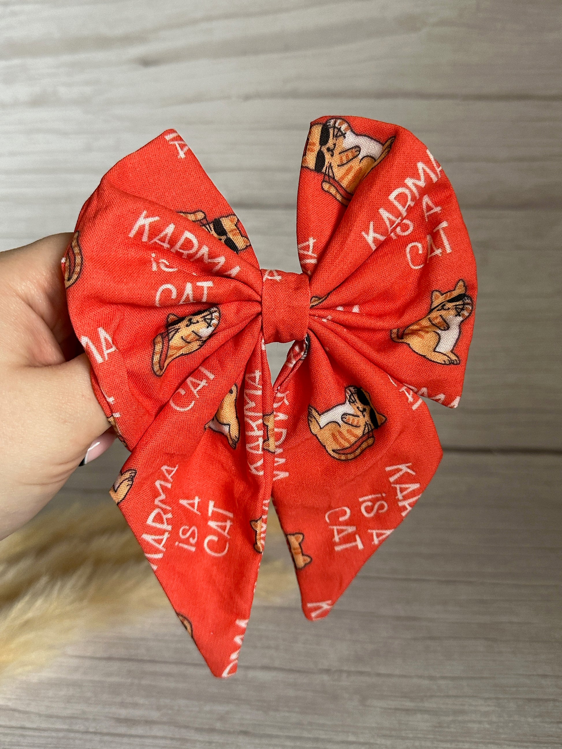 A hand holding the Cotton Sailor Bow Clip - Karma is a Cat from Crafts by Kate, a luxurious orange bow made of soft cotton adorned with illustrations of cats wearing sunglasses and the text "KARMA IS A CAT" printed repeatedly on the fabric. The background features a light wood surface.