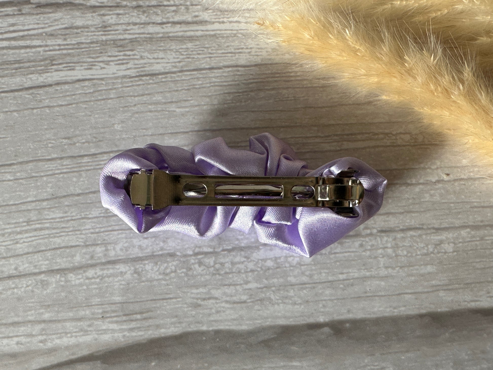 On a gray textured surface lies the elegant Silk French Barrette Scrunchie Clip in Lavender Purple by Crafts by Kate, while a furry beige object adds an intriguing touch from the top right corner.