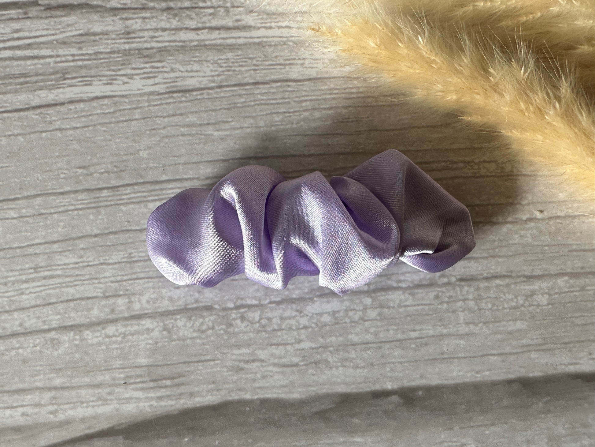 A luxurious Silk French Barrette Scrunchie Clip - Lavender Purple from Crafts by Kate rests on a textured gray surface, while a fluffy beige object peeks from the top right corner.