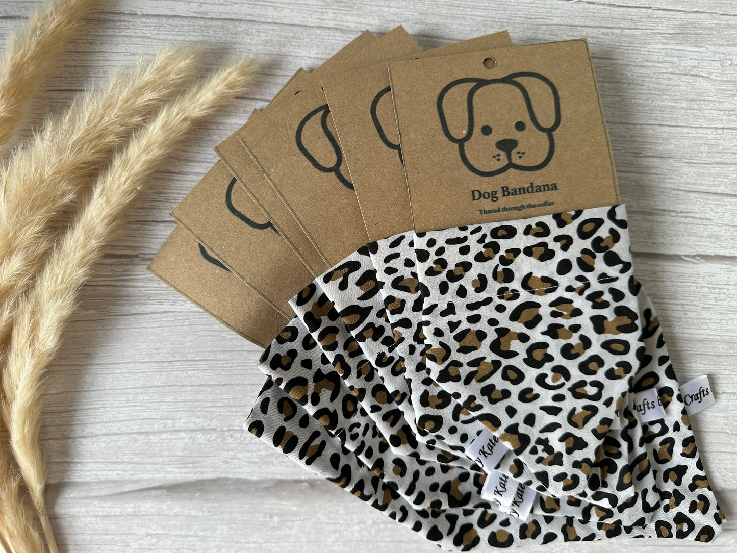 Several Cotton Pet Bandanas with collar attachments, designed in a leopard print by Crafts by Kate, are displayed on a light wooden surface. They are neatly packaged with brown cardboard tags featuring a simple illustration of a dog's face and labeled "Dog Bandana." Dried ornamental grass lies to the left.