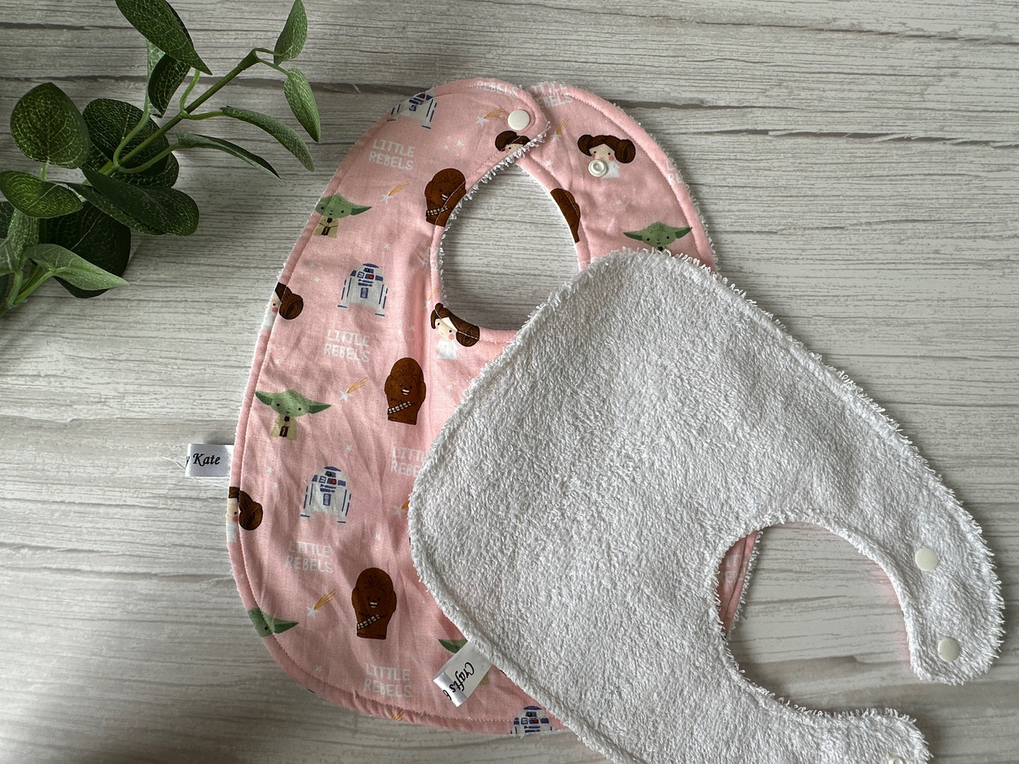 Two ultra-soft cotton baby bibs from the Crafts by Kate collection rest on a wooden surface. The top bib showcases the Star Wars Little Rebels design with playful character illustrations, while the plain white bib behind it offers contrast. A green plant partially visible on the left enhances the handcrafted display.