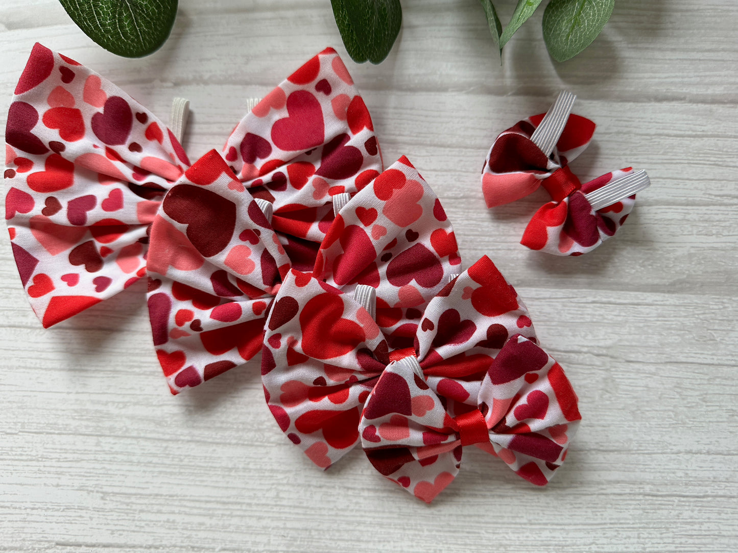 Four Crafts by Kate Cotton Pet Bows in Valentines Hearts fabric rest on wood. Three large, one smaller, all with elastic fastening for a snug fit. Green leaves gently add a hint of nature from above.