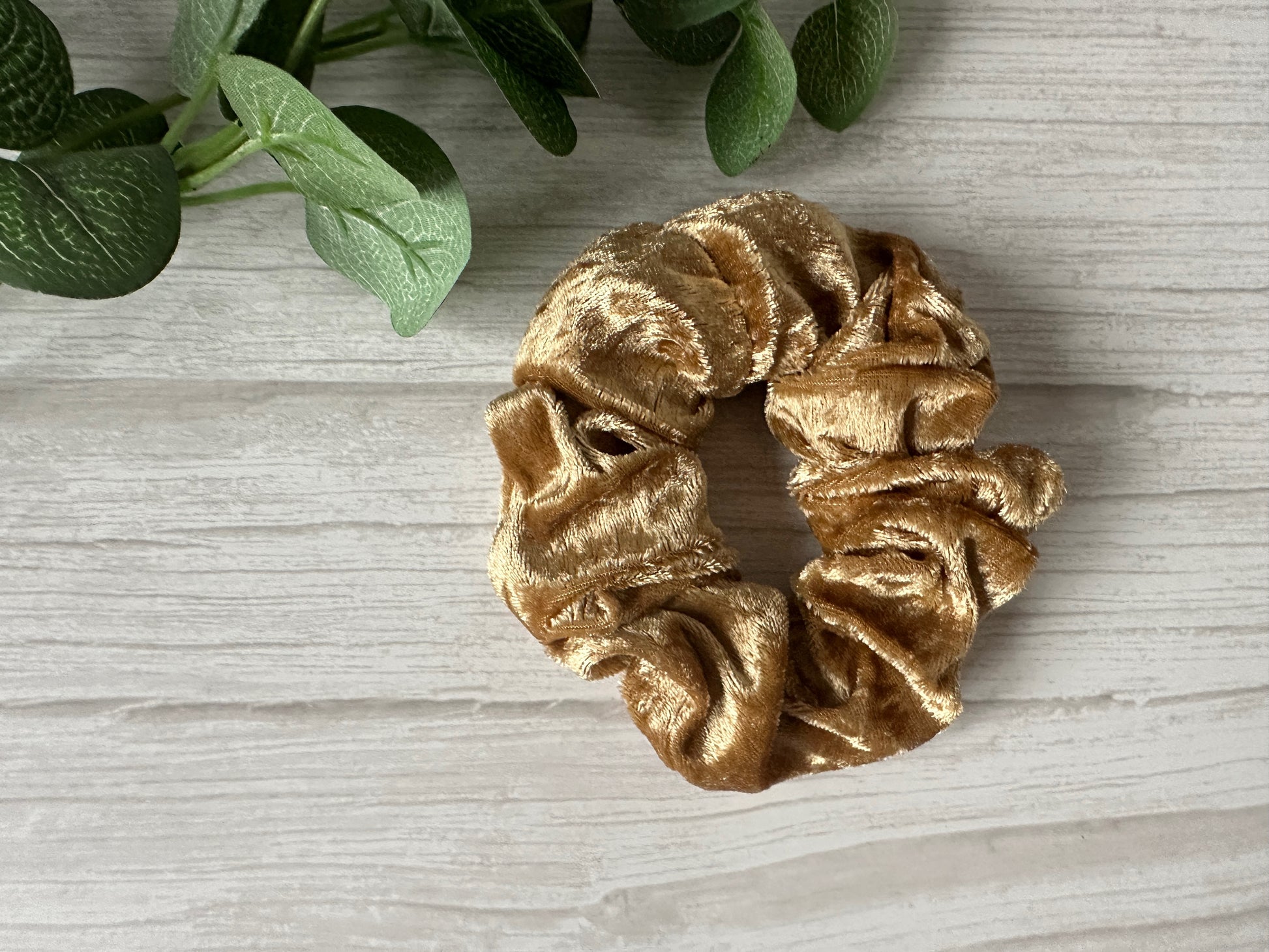 The Crushed Velvet Extra Large Scrunchie - Luxury Gold by Crafts by Kate sits on a light wooden surface, with green leaves in the corner, highlighting its luxurious golden sheen.