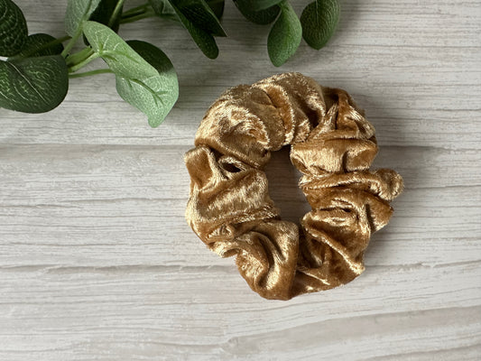 The Crushed Velvet Extra Large Scrunchie - Luxury Gold by Crafts by Kate sits on a light wooden surface, with green leaves in the corner, highlighting its luxurious golden sheen.
