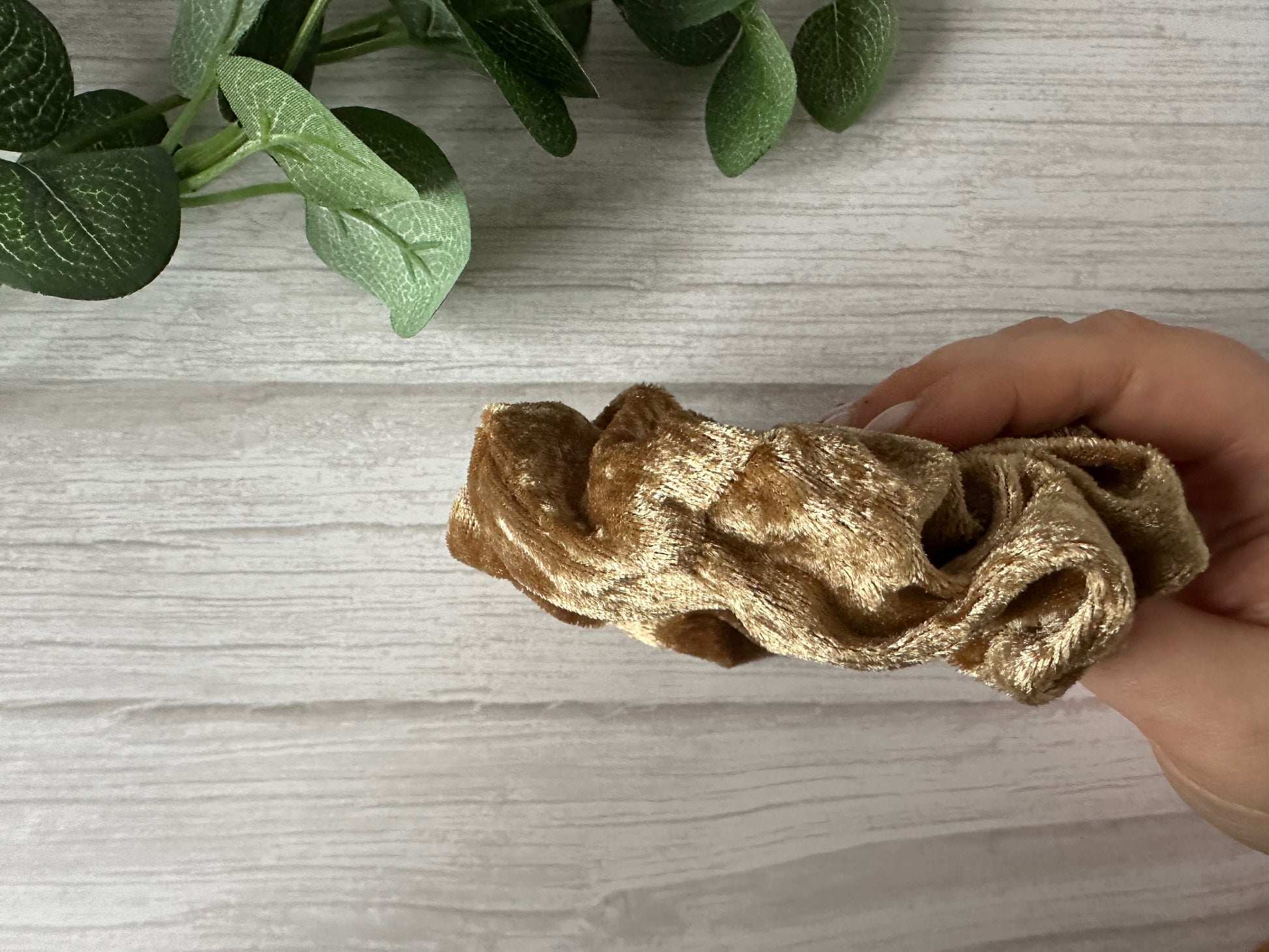 A hand holds the Crafts by Kate Crushed Velvet Extra Large Scrunchie in luxury gold over a light wooden surface, with green leaves visible in the top left corner.