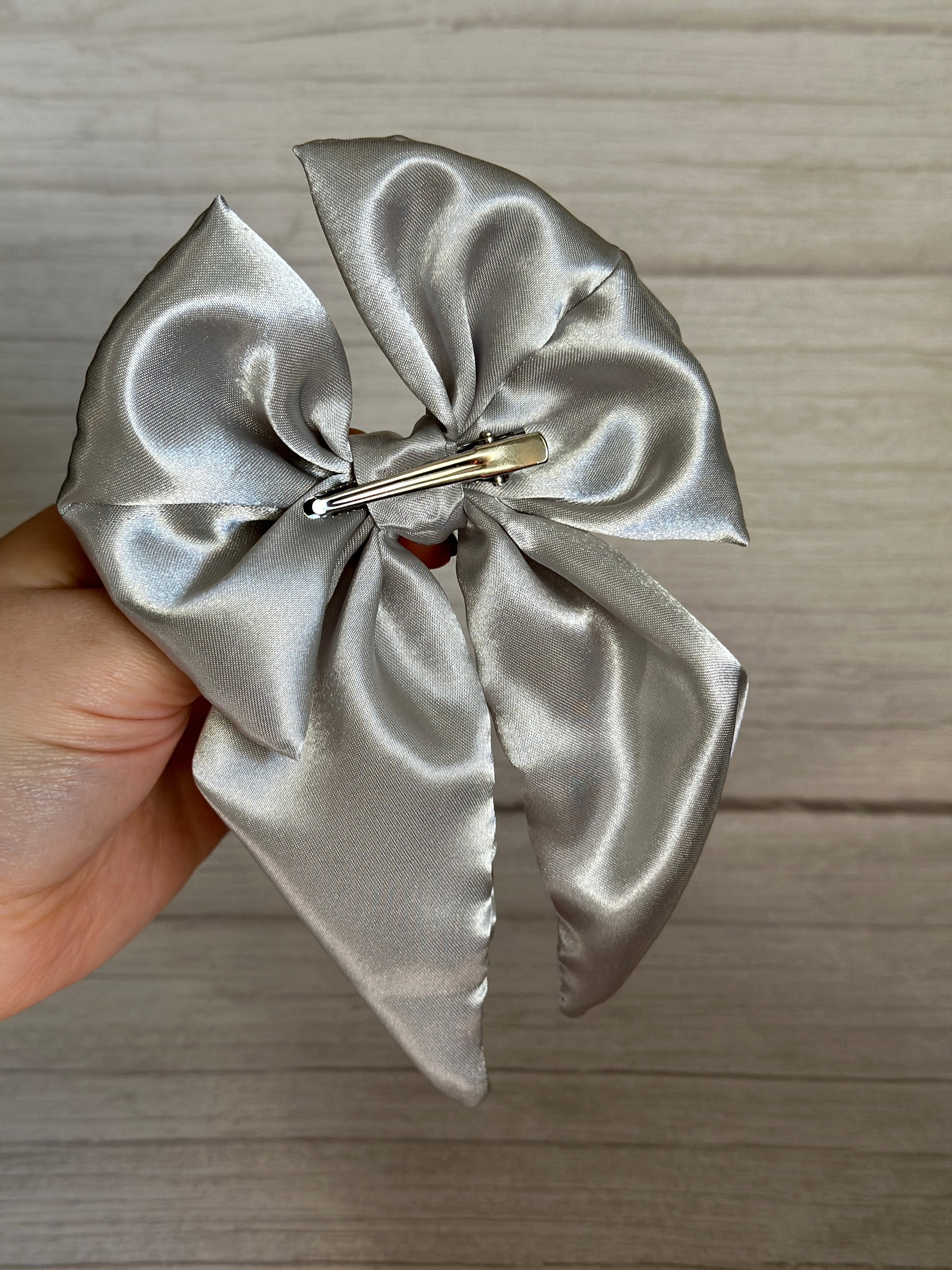 A hand holds the Silk Sailor Bow Clip - Metallic Silver by Crafts by Kate, showcasing its luxurious design. The handcrafted bow, made from silky smooth material with a shiny metallic silver finish, features elegant long tails and is attached to a hair clip. It rests gracefully against a light wooden surface.