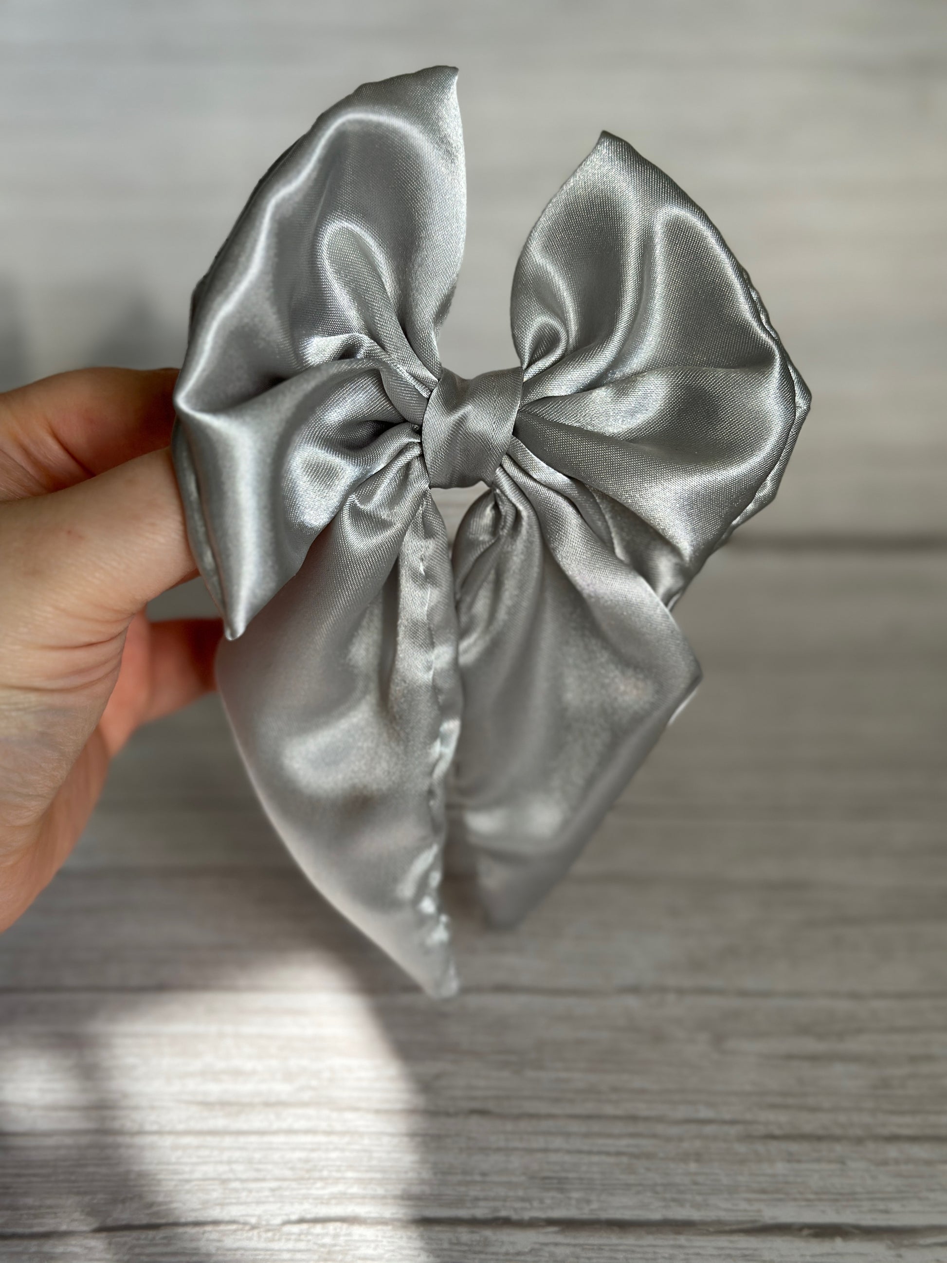 A hand displays the Crafts by Kate Silk Sailor Bow Clip in metallic silver, its silky texture glistening against the light gray wooden backdrop. The large bow, with its smooth and reflective surface, exudes an elegant and sophisticated vibe.