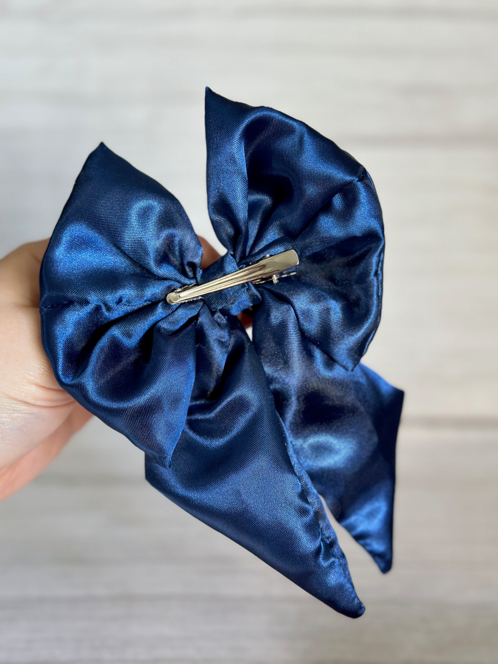 A hand holding the Silk Sailor Bow Clip in a luxurious, shiny midnight blue satin by Crafts by Kate, featuring a metal clip on the back. The bow showcases two large loops and long, trailing tails against a light, textured background.