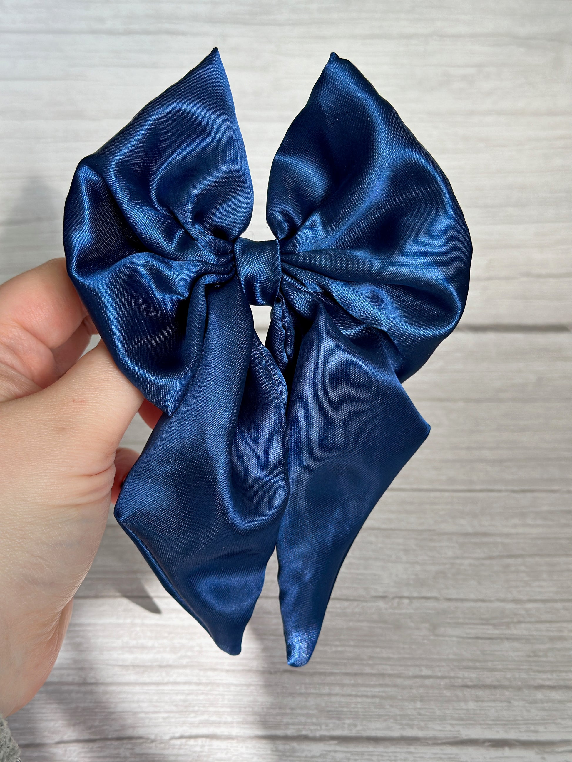 A hand elegantly presents the Silk Sailor Bow Clip in Midnight Blue by Crafts by Kate, its satin finish shimmering against a light wooden backdrop. The clip features symmetrical loops and long tails, offering a sophisticated and sleek look.