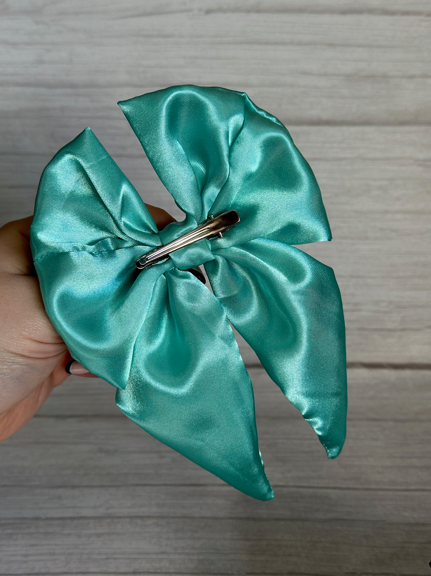 A hand holds the luxurious Silk Sailor Bow Clip - Mint Green from Crafts by Kate against a light gray background. The elegant bow, crafted from fine silk, features a metal hair clip on the back and two long tails.
