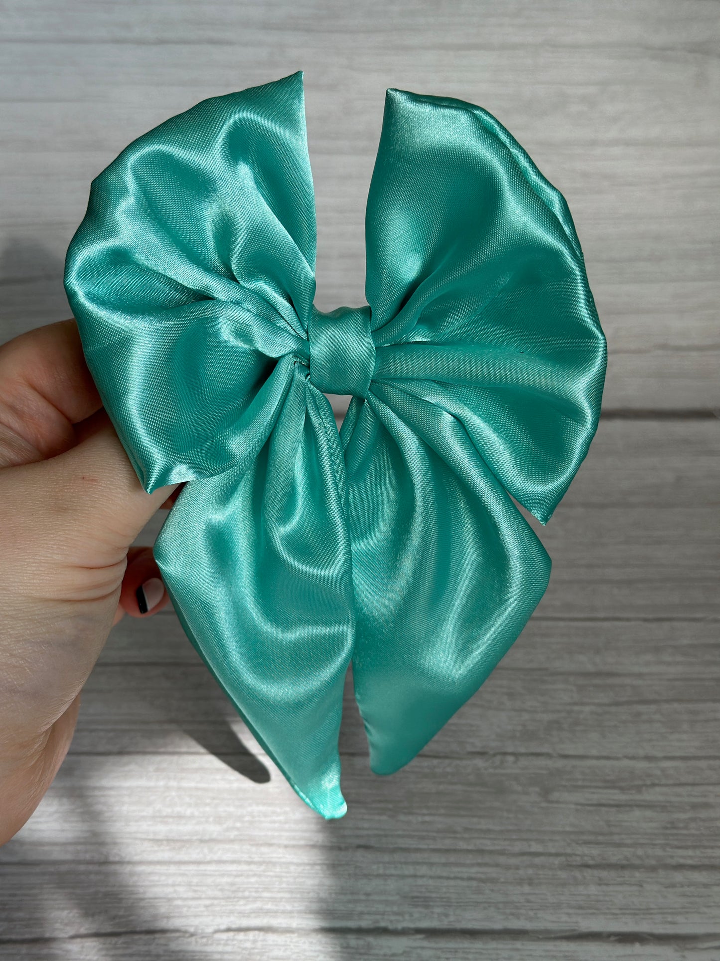 A hand elegantly holds the luxurious Silk Sailor Bow Clip in mint green by Crafts by Kate, featuring a shiny satin finish and a double-loop design, set against a light wood background.
