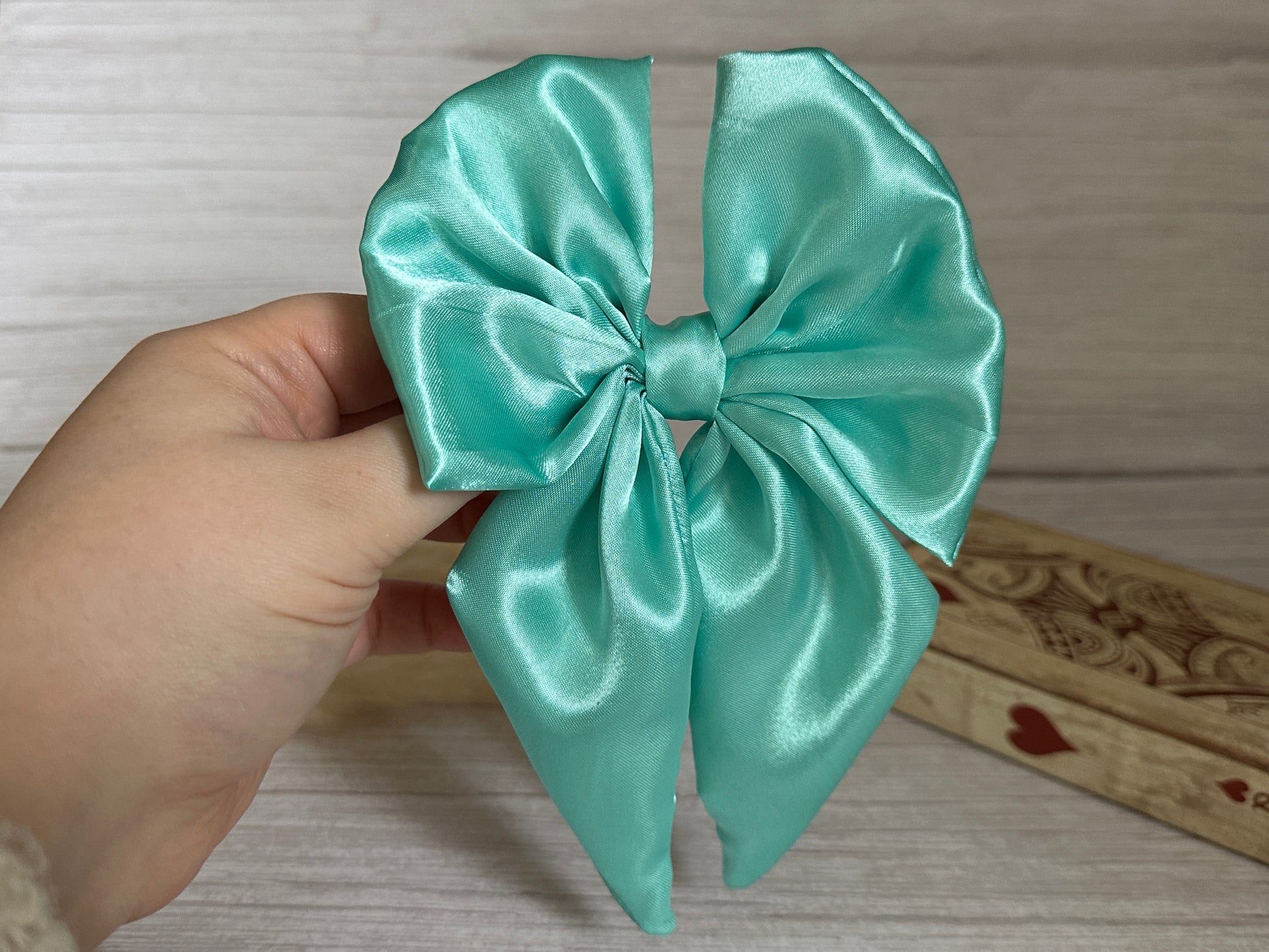 A hand holding the Silk Sailor Bow Clip in mint green from Crafts by Kate against a light wooden background. The bow has a silky texture and is neatly tied, with a decorative wooden box partially visible behind it.