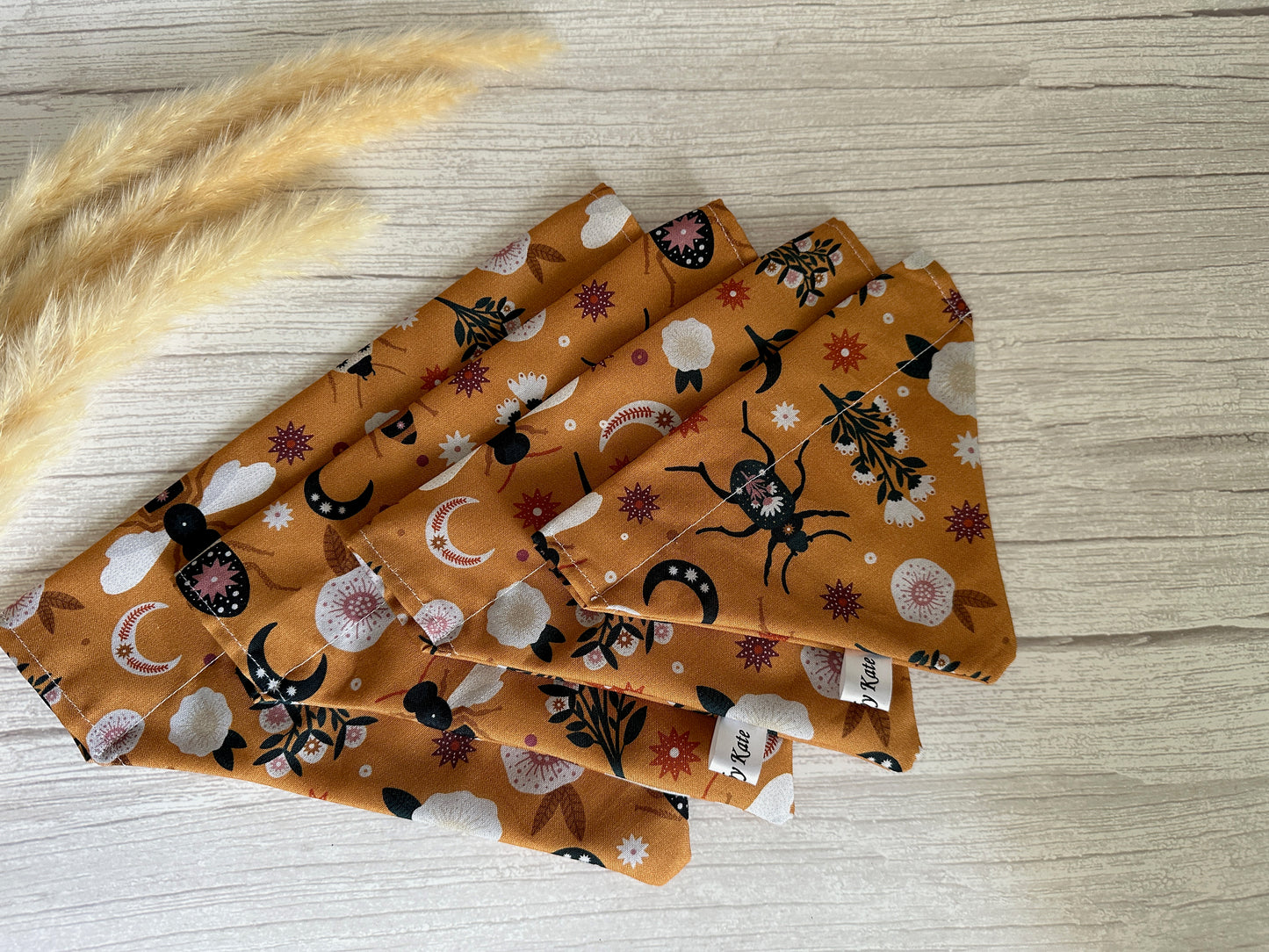 Four fabric napkins with autumnal motifs—leaves, acorns, moons, and flowers—are folded on a light wooden surface, accented by Crafts by Kate's Cotton Pet Bandana with collar attachment in orange floral, partially visible in the upper left corner alongside beige decorative grasses.