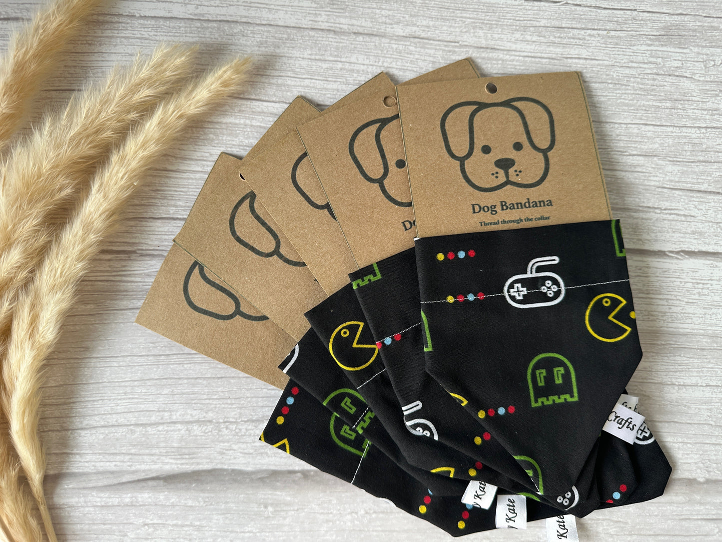 A set of five Cotton Pet Bandanas with collar attachments from Crafts by Kate, featuring retro gaming designs including a Pacman print fabric, are folded and arranged in a fan pattern. Each bandana comes with a kraft paper tag adorned with a simple dog illustration and the text "Dog Bandana." The bandanas and tags rest on a light wood surface next to dried wheat.