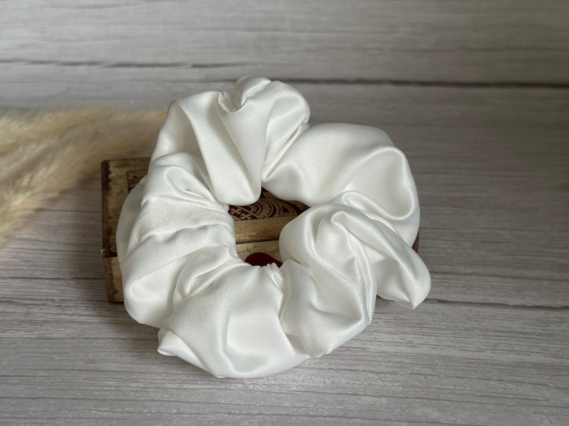 A handcrafted scrunchie from the Satin Extra Large Scrunchies - Bridal Collection by Crafts by Kate is elegantly displayed on a small, decorative wooden box. Its white satin shines beautifully against a neutral, textured background.