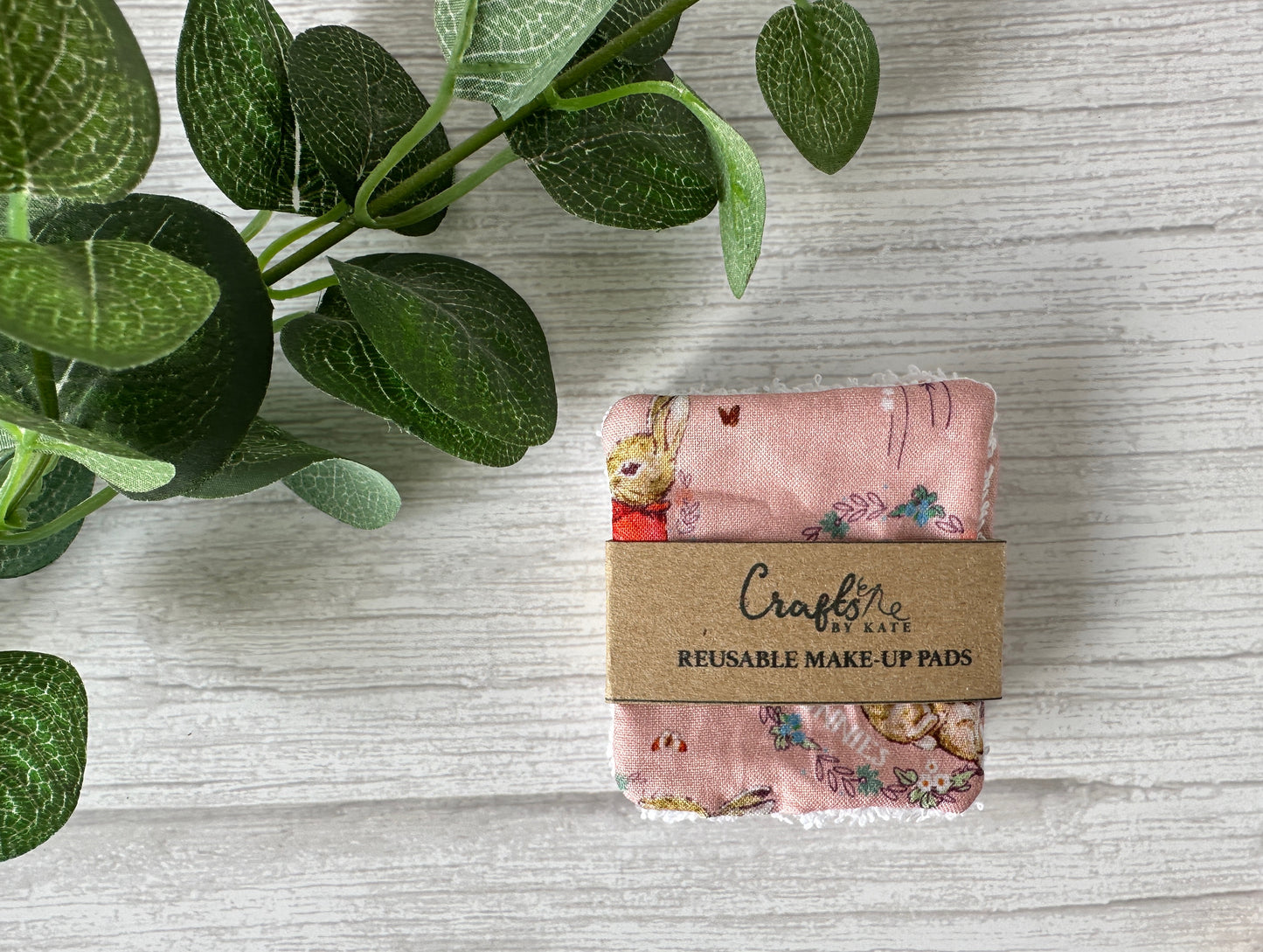 A set of sustainable Cotton Reusable Make-up Remover Pads - Peter Rabbit (pink) by Crafts by Kate, wrapped in a brown kraft paper label, rests on a light wooden surface next to green leaves.