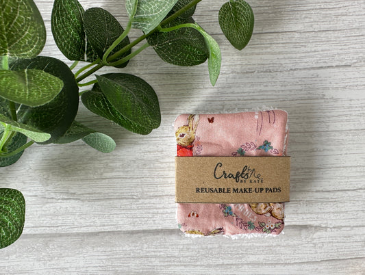 A set of sustainable Cotton Reusable Make-up Remover Pads - Peter Rabbit (pink) by Crafts by Kate, wrapped in a brown kraft paper label, rests on a light wooden surface next to green leaves.