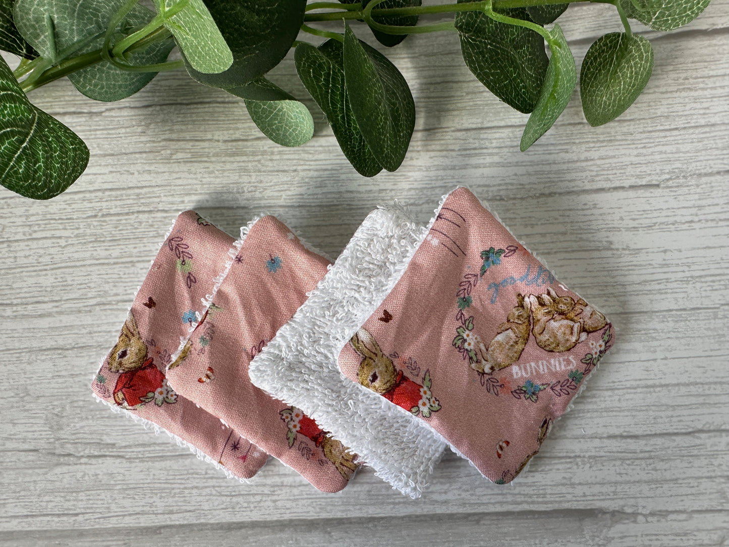 Four Crafts by Kate Cotton Reusable Make-up Remover Pads - Peter Rabbit (pink), featuring a bunny pattern and soft white side, are stacked on light wood. Green leaves add a natural touch in the top left corner of the image.