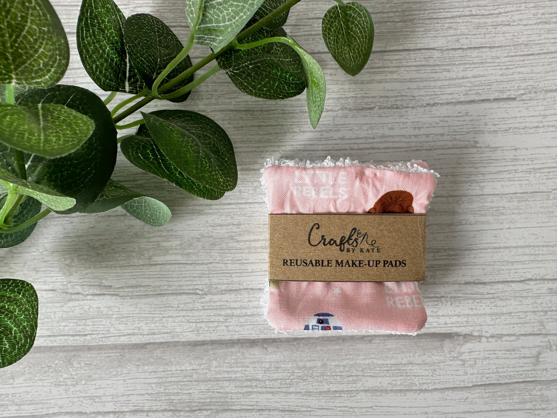 A neatly folded stack of sustainable Crafts by Kate Cotton Reusable Make-up Remover Pads - Star Wars Little Rebel, on a wooden surface. A green plant with round leaves in the upper left corner enhances the eco-friendly aesthetic.