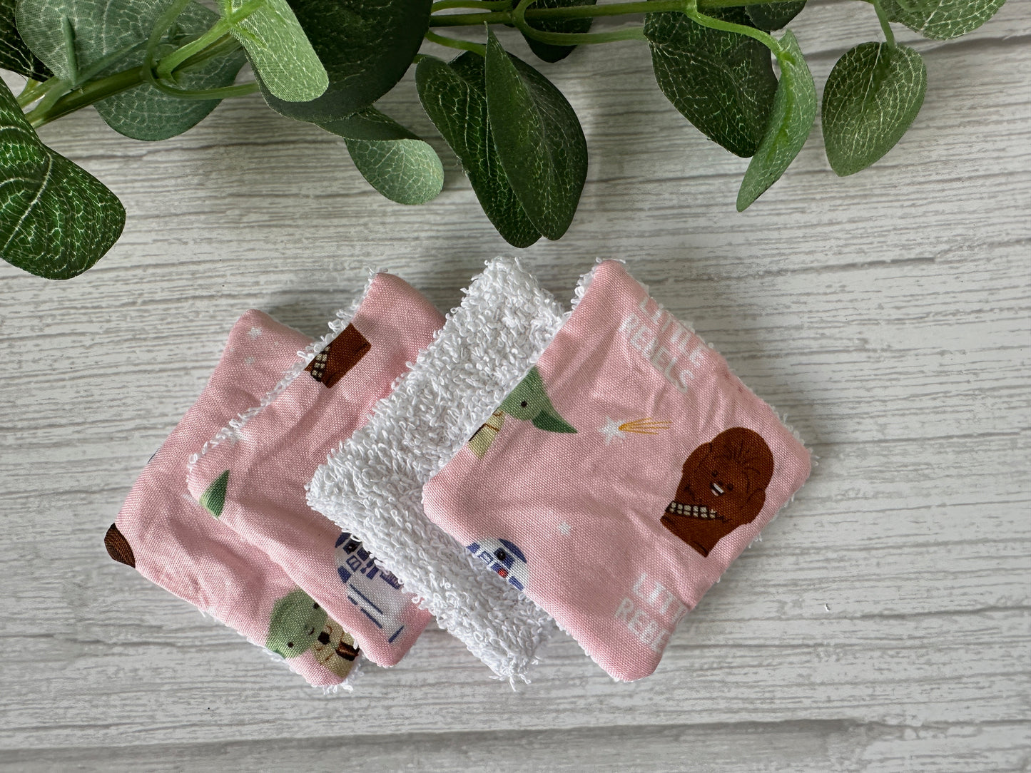 On a light gray wooden surface, three eco-friendly Crafts by Kate Cotton Reusable Make-up Remover Pads - Star Wars Little Rebel show characters like Yoda and Chewbacca, with a leafy green plant partially visible in the upper left corner.