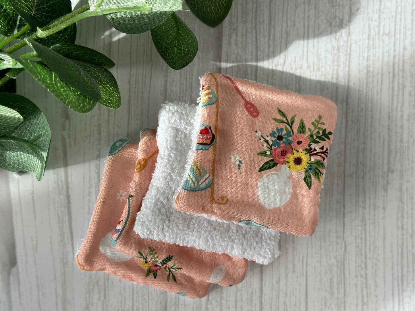 Four pink floral patterned Cotton Reusable Make-up Remover Pads by Crafts by Kate are neatly arranged on a light wood surface. A green plant leaf casts a shadow in the top left corner, emphasizing sustainability.
