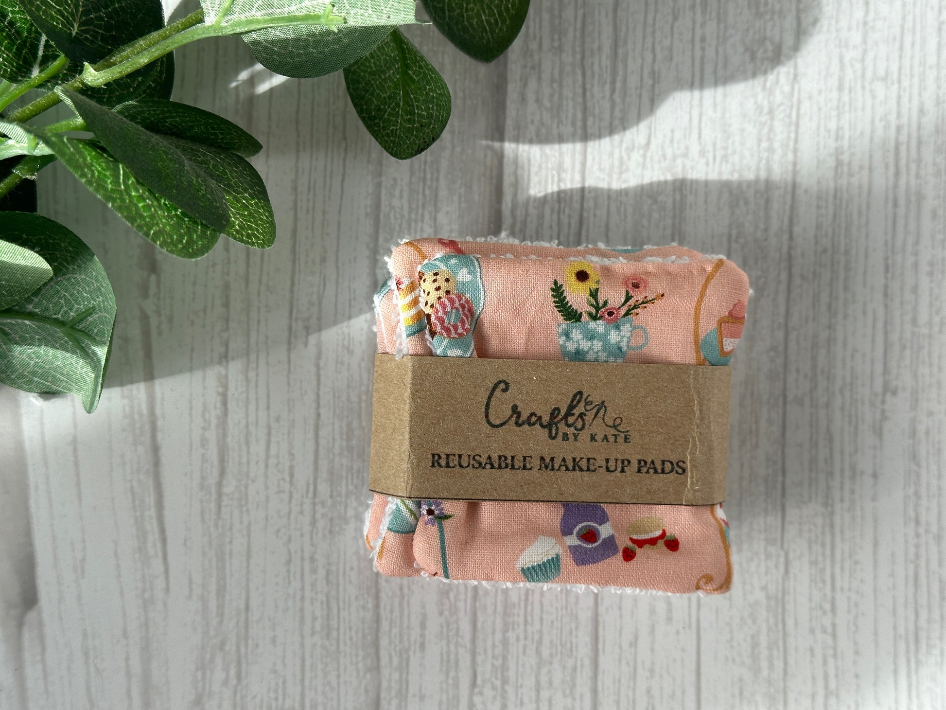 A set of Cotton Reusable Make-up Remover Pads by Crafts by Kate, featuring the "Pink Tearoom" design, sits on a light wooden surface. The pads showcase a vibrant pattern with makeup items and are wrapped in a paper band labeled "Crochet." A green leafy plant adorns the top left, enhancing their sustainable appeal.