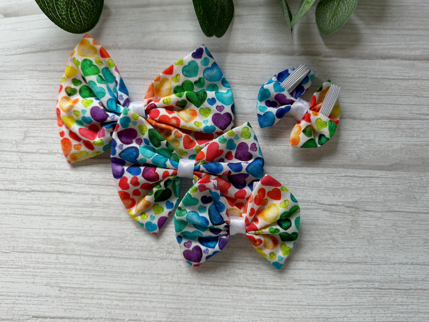 Crafts by Kate's Cotton Pet Bow with collar attachment - Rainbow Hearts, elegantly displayed on a light wooden surface, is accented by the natural touch of green leaves partially visible in the corner.
