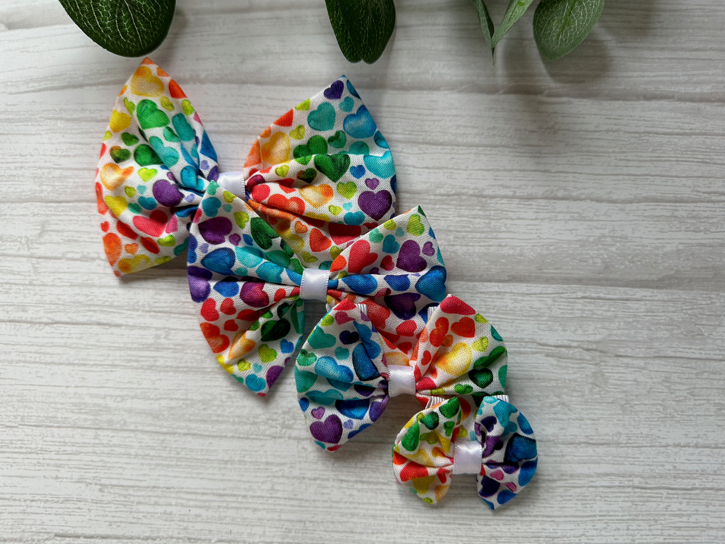 Three vibrant Cotton Pet Bows with collar attachments from Crafts by Kate, made with Rainbow Hearts fabric, are laid out in descending size on a light wooden surface. A hint of greenery peeks in at the top left, adding a natural touch to the display.