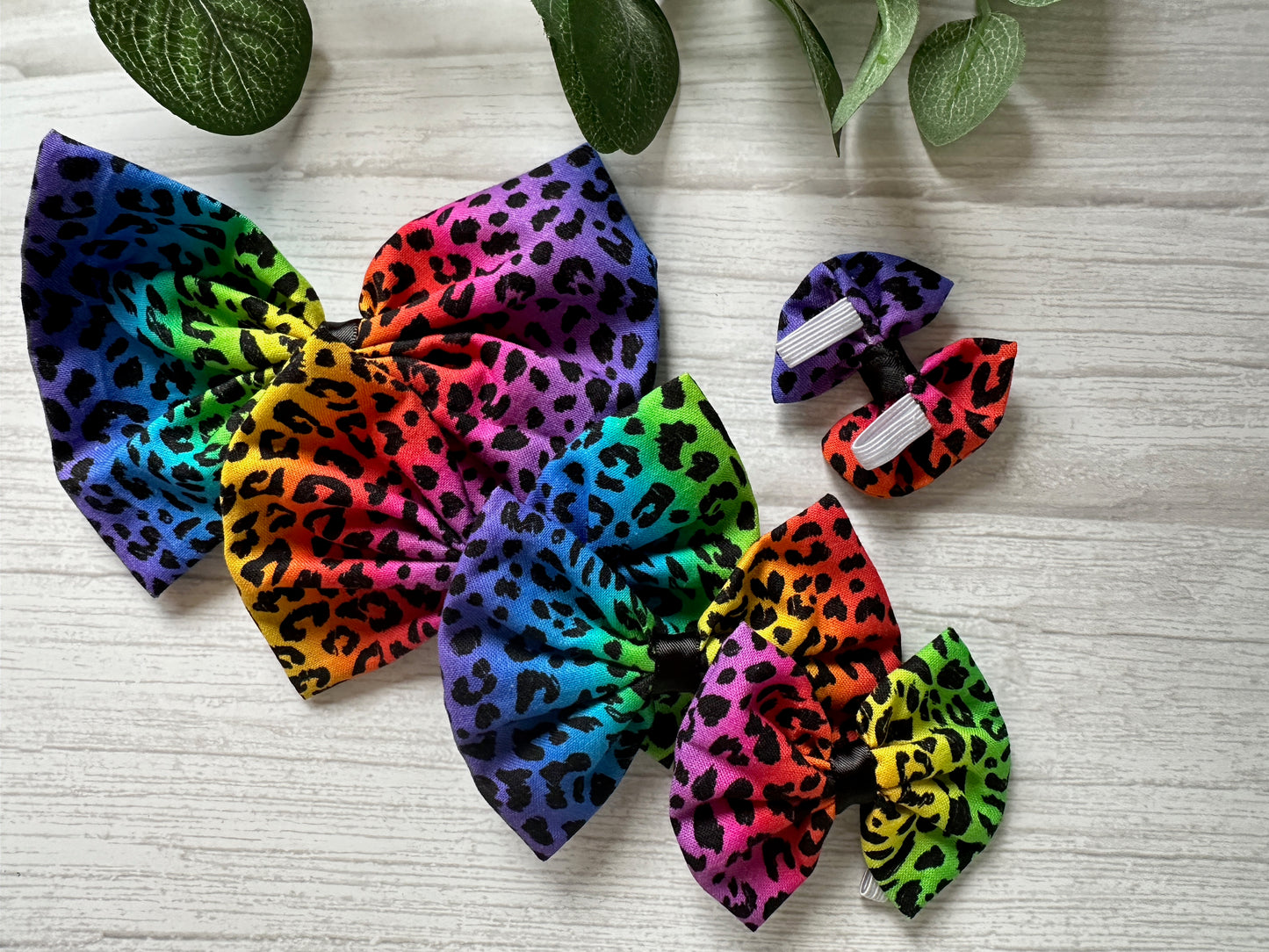On a white wooden surface, Cotton Pet Bows by Crafts by Kate, featuring rainbow leopard print and collar attachments, are artfully displayed with green leaves adding a natural touch.