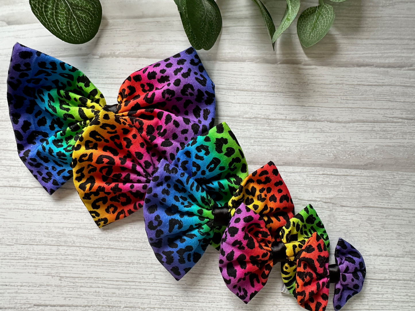 The Cotton Pet Bow set by Crafts by Kate includes four vibrantly colored rainbow leopard print bows in decreasing sizes, designed for collar attachment, elegantly displayed on a light wooden surface with green leaves accentuating the top corners.