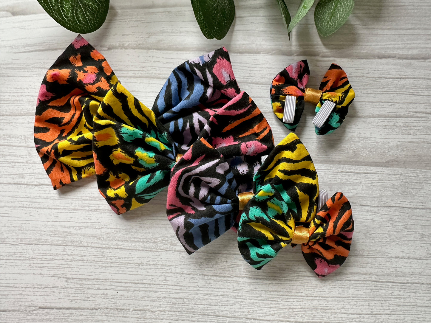 Four colorful Cotton Pet Bows by Crafts by Kate, made from Rainbow Tiger Print Fabric with collar attachments, are arranged on a light wooden surface. Their playful sizes contrast with the green leaves peeking into the image.