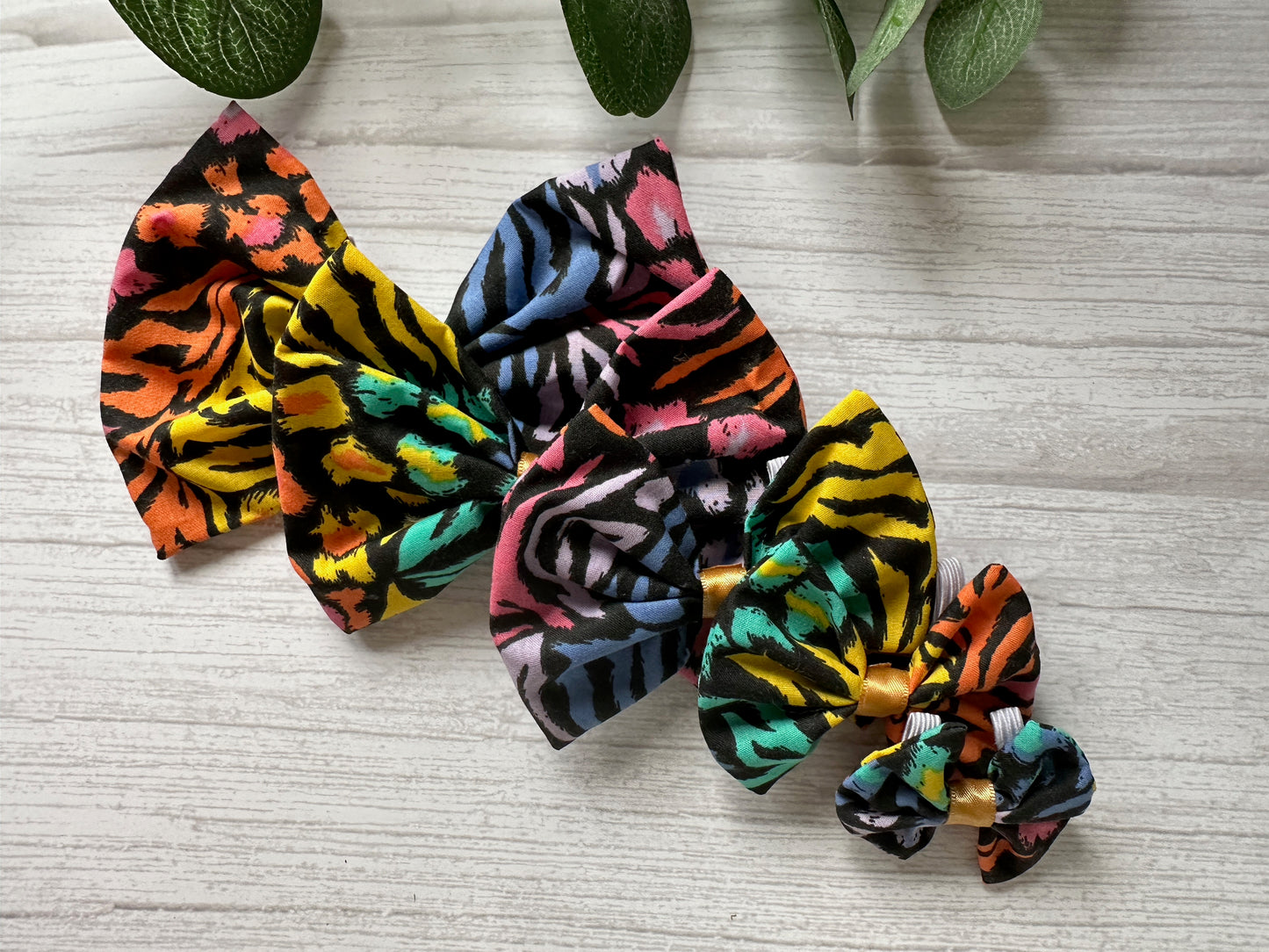 Four Crafts by Kate Cotton Pet Bows in vibrant Rainbow Tiger Print (yellow, pink, blue, green, and orange) sit on light wood. The elastic attachments make them pup-ready. Green leaves peek from the top.