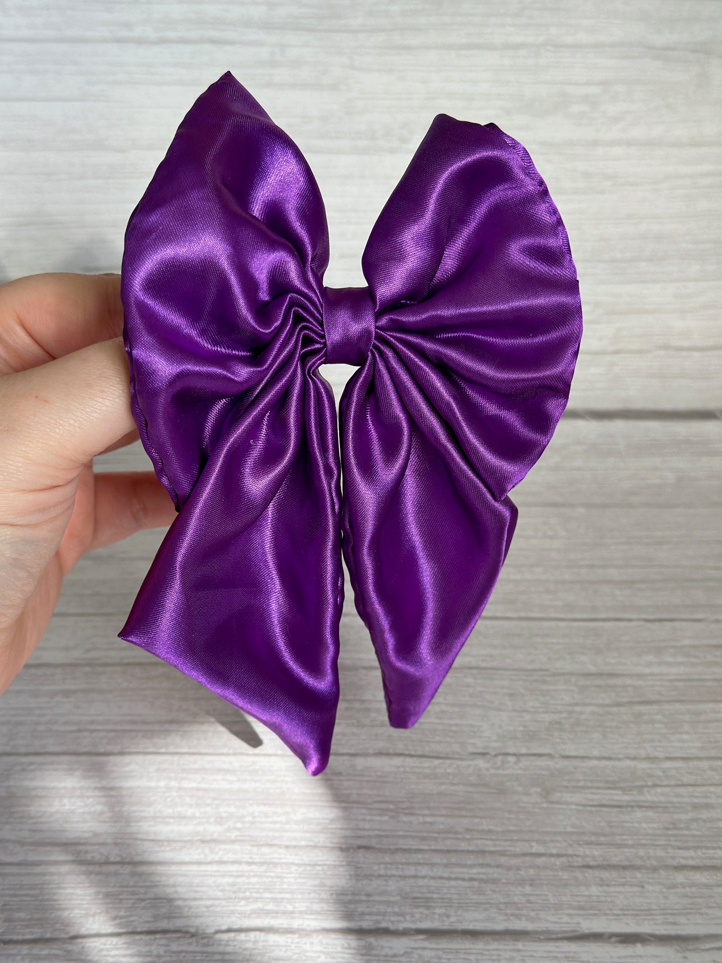 A hand holds the Silk Sailor Bow Clip - Royal Purple by Crafts by Kate against a light wooden background. The large, symmetrical bow resembles a statement sailor bow with gathered fabric in the center, creating an elegant ruffled texture.
