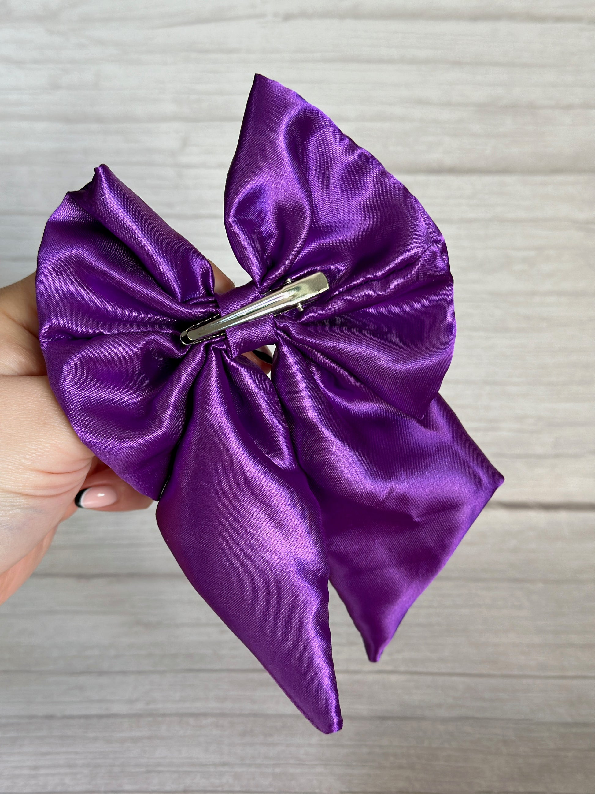 A hand holds the elegant *Silk Sailor Bow Clip - Royal Purple* by Crafts by Kate, featuring a luxurious satin bow attached to a sleek silver hair clip, creating a perfect statement accessory. The background is a light gray textured surface.