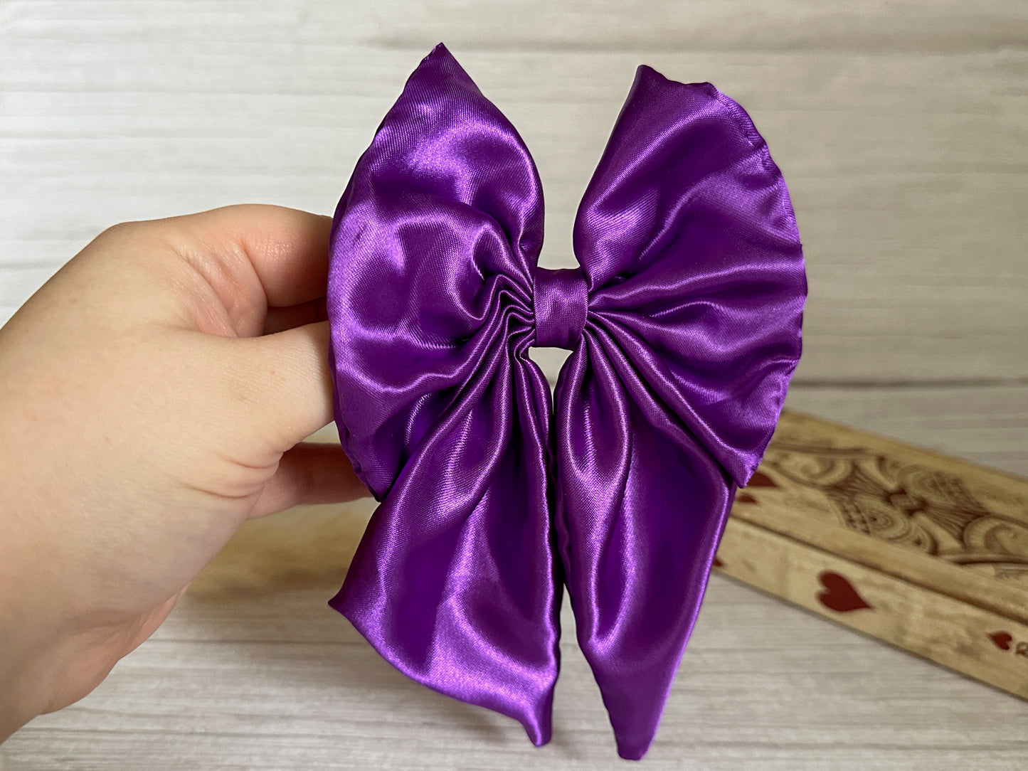 A hand holds the Crafts by Kate "Silk Sailor Bow Clip" in royal purple against a light wooden background. Behind the bow, a rectangular piece of wood adorned with heart decorations is partially visible, creating an elegant and charming scene.