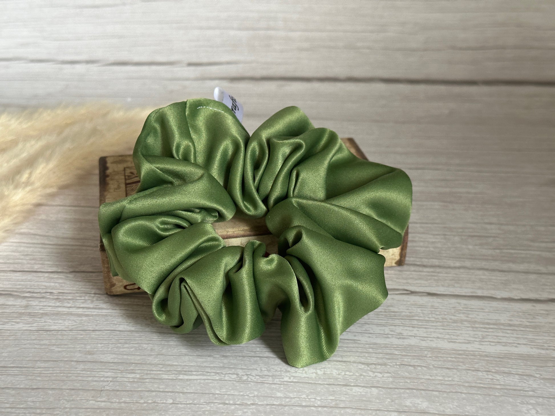 A Satin Extra Large Scrunchie from the Bridal Collection by Crafts by Kate rests atop a wooden box on a light wood surface. This handcrafted statement piece, in a soothing green hue, is gathered in soft folds that create a textured appearance. To the left, a small tuft of beige pampas grass partially peeks into view.
