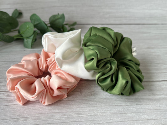 A trio of satin extra-large scrunchies from the Crafts by Kate Bridal Collection, available in pastel pink, white, and olive green shades, are artfully displayed on a light wooden surface. A small green plant decorates the upper left corner of the image, enhancing the natural elegance of this luxurious collection.