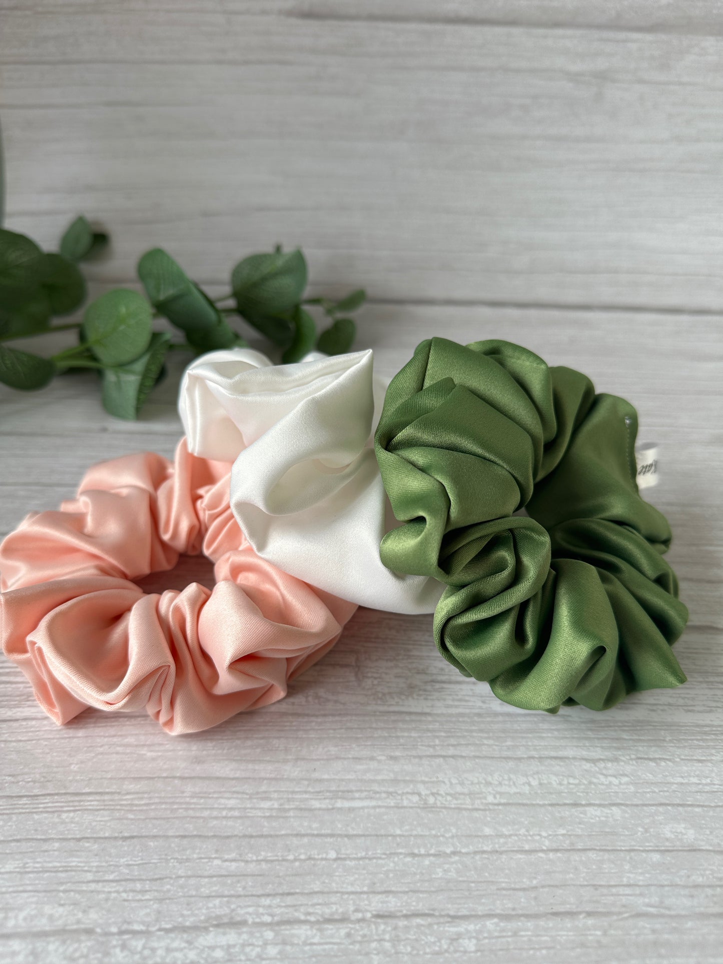 Three satin extra large scrunchies from the Bridal Collection by Crafts by Kate, in shades of pink, white, and green, are elegantly displayed on a light wooden surface. A green leafy branch in the background enhances the natural elegance of this handcrafted statement piece.