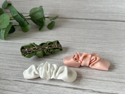 Displayed on a light wooden surface is a set of three satin fabric hair bows from our Crafts by Kate Bridal Collection, available in white, light pink, and green. The green bow, known as the Satin Scrunchie Style French Barrette - Bridal Collection, is shown with its metal clasp facing up. A sprig of green leaves lies in the background.