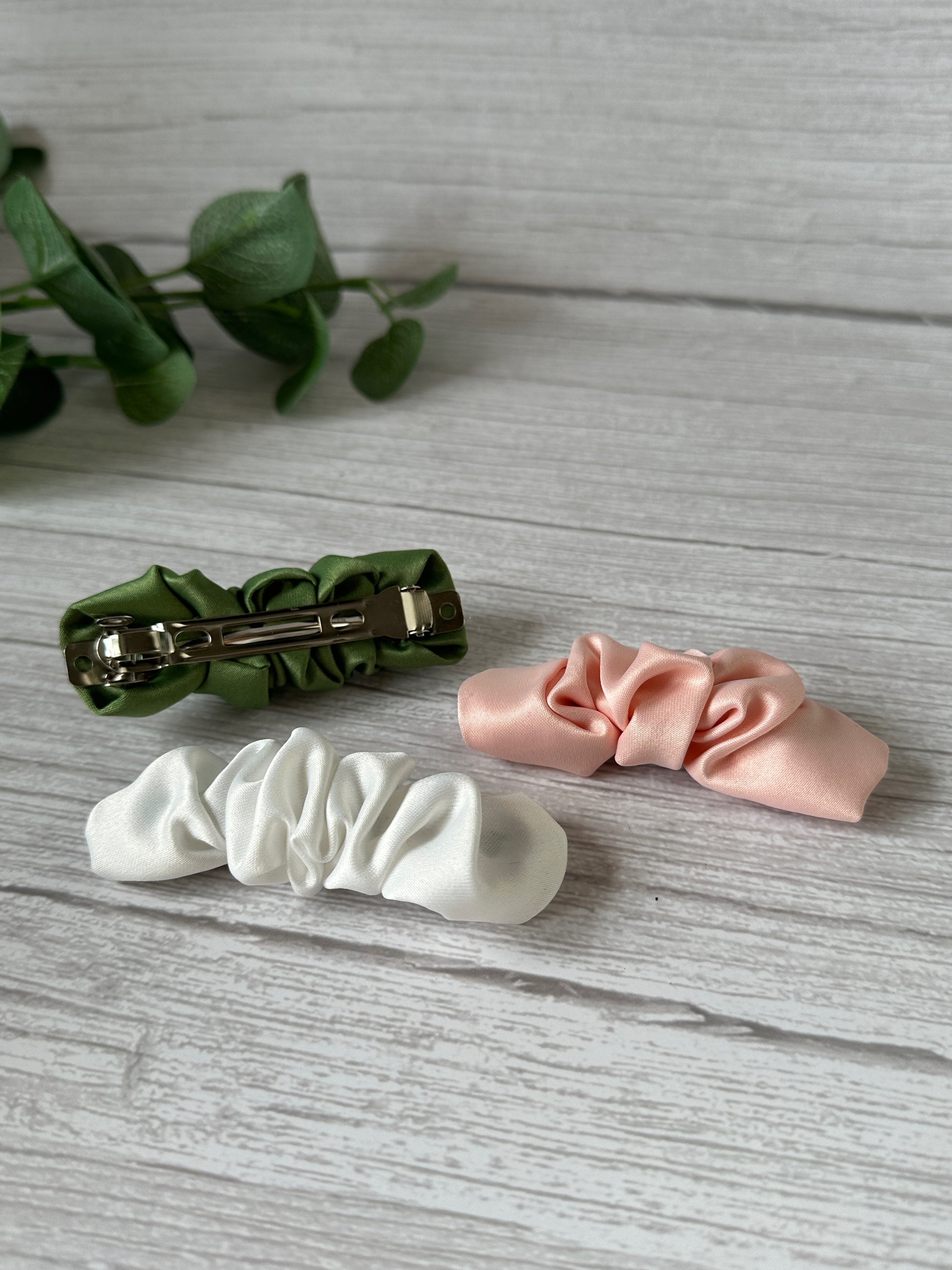 Image of three Satin Scrunchie Style French Barrettes from the Bridal Collection by Crafts by Kate in green, white, and pink, placed on a white wood surface. Two barrettes are arranged face up, while the green one is face down, partially revealing its gold French barrette closure. A small green plant is in the background.