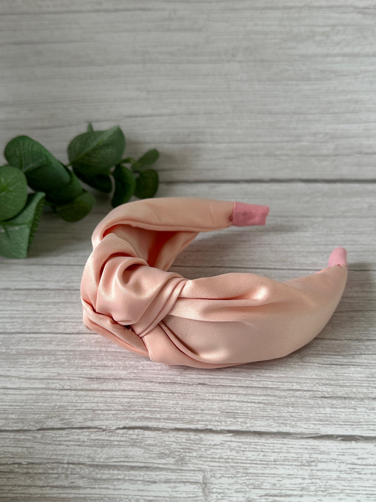 A soft pink Satin Knotted Headband from Crafts by Kate's Bridal Collection is elegantly displayed on a light wooden surface, with a knotted design. A small green plant in the background adds a vibrant touch of color.