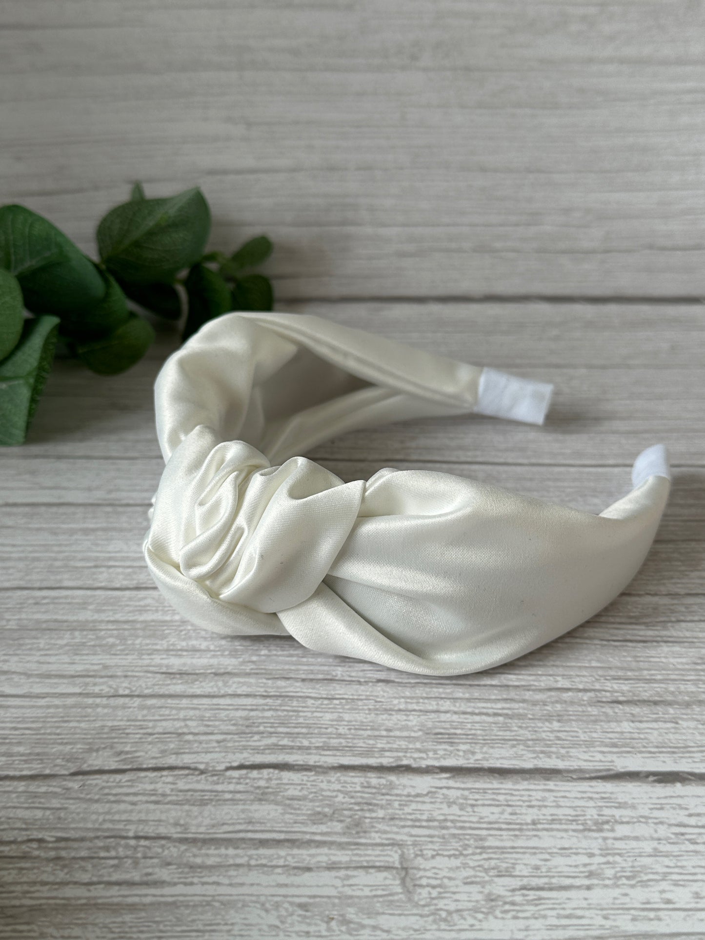 A Satin Knotted Headband from the Bridal Collection by Crafts by Kate rests on a light wooden surface. The headband features a large twisted knot, and green leaves in the background add a touch of nature to the scene. This elegant piece boasts a cream color and smooth texture.