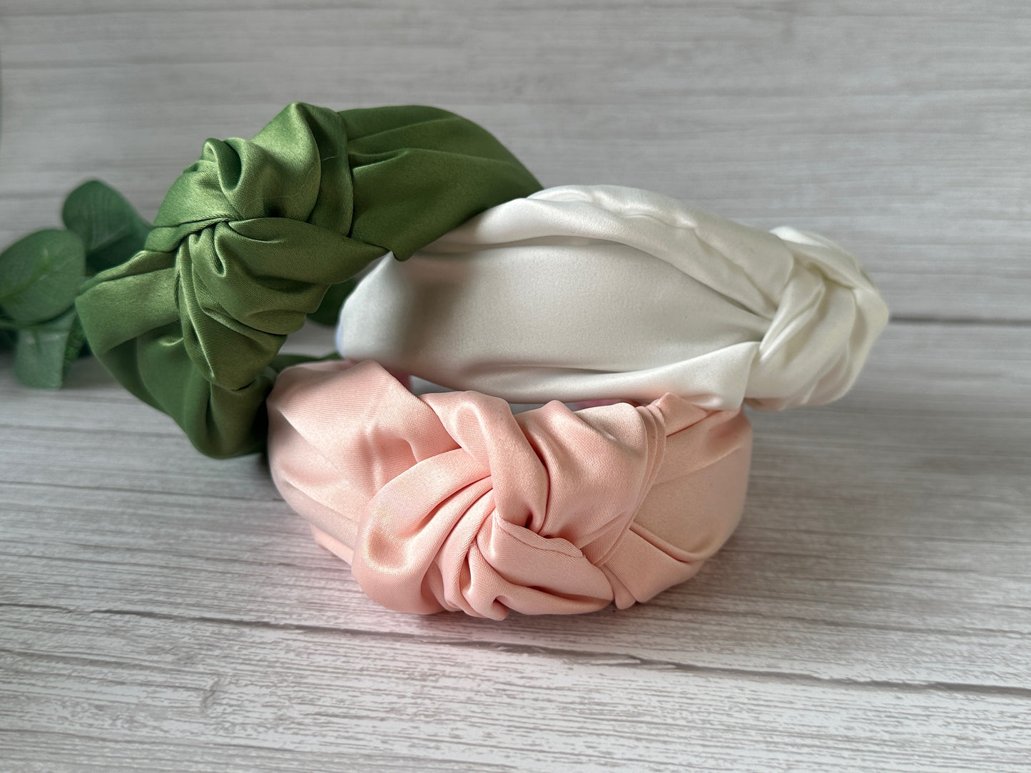 Three Satin Knotted Headbands from the Crafts by Kate Bridal Collection are elegantly showcased on a wooden surface. These handcrafted headbands, available in green, white, and pink, feature a shiny, smooth texture and are artfully arranged on top of each other, making them an ideal addition to any bridal collection.