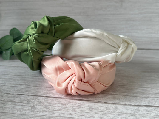Three Satin Knotted Headbands from the Crafts by Kate Bridal Collection are elegantly showcased on a wooden surface. These handcrafted headbands, available in green, white, and pink, feature a shiny, smooth texture and are artfully arranged on top of each other, making them an ideal addition to any bridal collection.