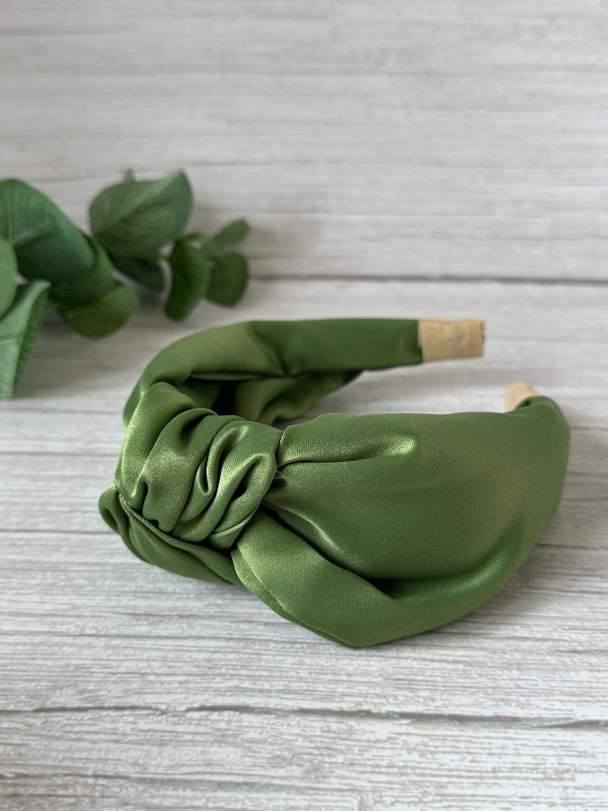 A Satin Knotted Headband from the Bridal Collection by Crafts by Kate, featuring a handcrafted green satin design, is elegantly displayed on a light wooden surface. In the background, a small sprig of eucalyptus enhances its exquisite charm.